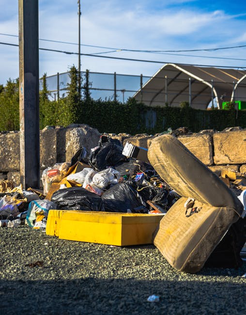 An outdoor scene showing a pile of mixed waste and rubbish, including black plastic trash bags, cardboard boxes, and loose debris, situated on a gravel surface next to a low stone wall made of large, rough-hewn stones. A large, weathered beige tyre leans against the rubbish heap, partially covering some of the waste. Behind the wall, there is a green hedge and a clear blue sky with a few clouds, while overhead power lines run horizontally across the background. To the left, a tall metal utility pole is visible. The scene likely depicts an area awaiting collection by private waste removal services such as Waste Clearance Kingston, emphasizing the importance of professional rubbish disposal and alternative waste handling outside of municipal services.