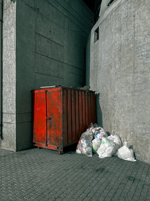 A large, rusty red metal skip with vertical ridges is positioned against a dark green wall and a light grey concrete wall in an urban outdoor area. Several white plastic bags filled with various types of rubbish are stacked next to and leaning against the skip, containing what appear to be general waste and recyclables. The skip sits on textured paving stones on the ground, which extend into the environment. In the background, a small window is visible high on the dark green wall. The scene is illuminated by natural light, casting soft shadows, and suggests an environment where waste collection, possibly an alternative to council services, is taking place. The presence of Waste Clearance Kingston indicates a professional private waste removal service providing on-site clearance or independent collection in this setting.