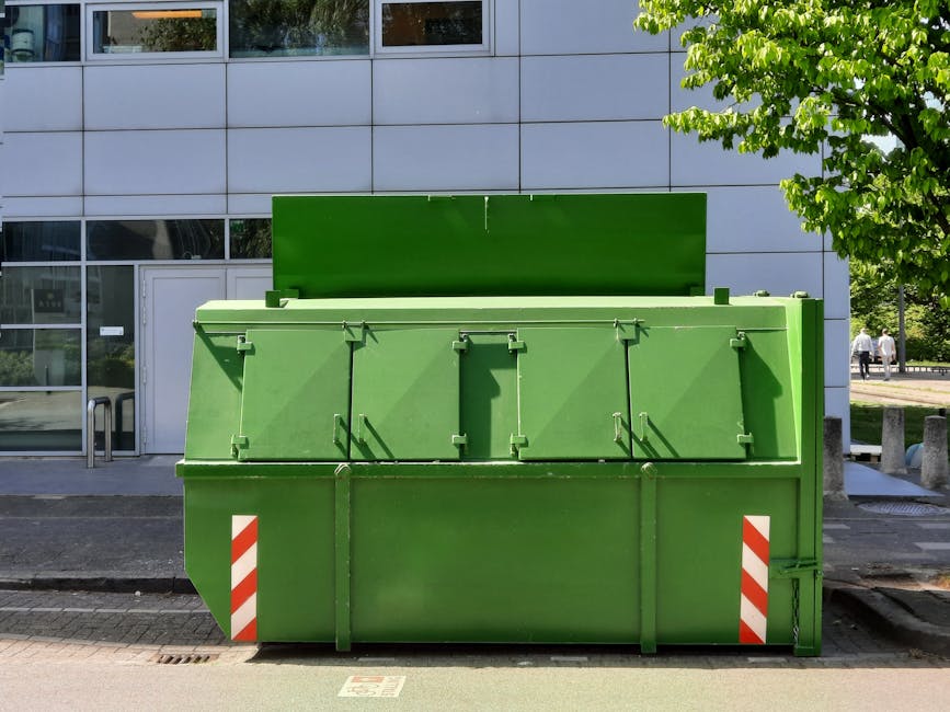 A large, rectangular, bright green waste container is positioned on a paved sidewalk in front of a modern building with a grey tiled facade and large glass windows. The container has a matte finish with visible hinges and locking mechanisms on its front panel, which features a slightly curved top edge and is divided into several sections by vertical metal bars. The lower part of the container has diagonal red and white reflective strips on each corner for visibility and safety. The environment surrounding the container includes a smooth asphalt road, a nearby white door to the building, and a small tree with vibrant green leaves on the right side, providing some natural contrast against the urban setting. Two pedestrians can be seen in the distance on the sidewalk, and the scene is well-lit with natural daylight, casting shadows from the container onto the pavement. This scene likely depicts an independent waste collection point, consistent with private rubbish management services in an urban environment, with Waste Clearance Kingston operating in the background of such urban waste disposal solutions.