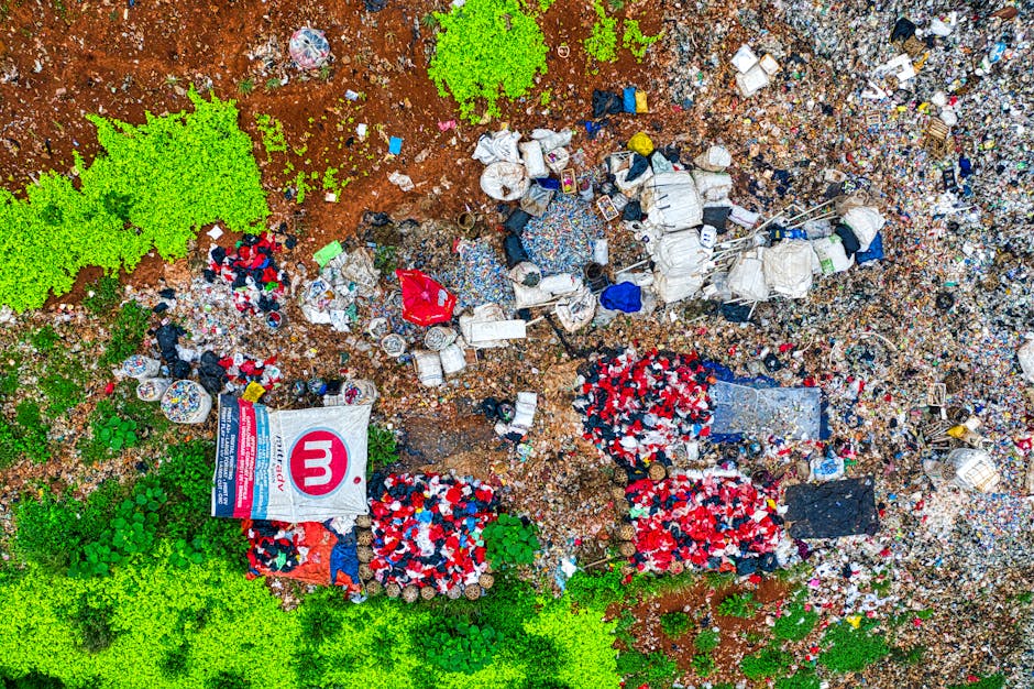 An aerial view of a large, unorganized landfill site featuring a mixture of waste materials spread across uneven ground. In the foreground, there are bright green patches of foliage and small trees that border the waste area, providing a stark contrast to the surrounding rubbish. The landfill contains numerous plastic debris, such as bottles, bags, and containers, along with larger items like rolled-up carpets, cardboard, wooden pallets, and discarded furniture. Some areas include clusters of red, white, and blue discarded ball-shaped items, possibly packaging or protective padding. A prominent white bag with a red 'm' logo and a blue and red stripe design is visible near the bottom left, lying partially open among the waste. The overall scene depicts a mixture of raw, textured materials in various colors, with a sense of clutter and disorder, typical of unmanaged waste dumps. The background extends further with densely piled debris, emphasizing an environment affected by private or independent waste disposal methods, rather than systematic collection.