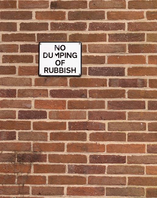 A white rectangular sign with black lettering mounted on a red brick wall reads 'NO DUMPING OF RUBBISH' in bold, uppercase text. The sign is positioned centrally in the upper-left section of the image, with the brick wall extending horizontally and vertically around it. The bricks have a textured surface with various shades of red, brown, and occasional darker hues, creating a traditional brickwork pattern with mortar joints visible between each brick. The lighting appears natural, evenly illuminating the wall and sign without harsh shadows. This scene depicts a clear warning against illegal rubbish dumping at a residential or commercial property, consistent with private waste management regulations often handled by services such as Waste Clearance Kingston, who offer alternatives to traditional local authority collection for rubbish removal.