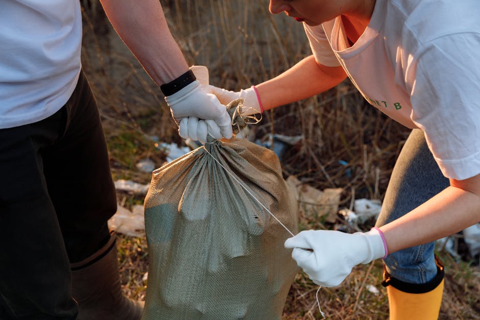 Two individuals wearing white gloves are engaged in rubbish collection outdoors, with one person holding a tan fabric sack filled with waste, while the other secures it with a drawstring. They are dressed in casual clothing, with visible sleeves and partial torsos, working in a natural setting with dry grass, twigs, and scattered debris on the ground. The background suggests an overgrown or neglected area, possibly a verge or vacant lot, where informal waste disposal or fly-tipping might occur. The scene emphasizes private or independent rubbish removal efforts, aligning with services such as those offered by waste clearance companies like Waste Clearance Kingston, which specialise in rubbish collection and site clearance tasks. The lighting appears natural, indicative of daylight, highlighting the textures of the waste material and the fabric of the sack, contributing to a clear, precise visual description suitable for accessibility and SEO relevance concerning waste management and rubbish removal services.