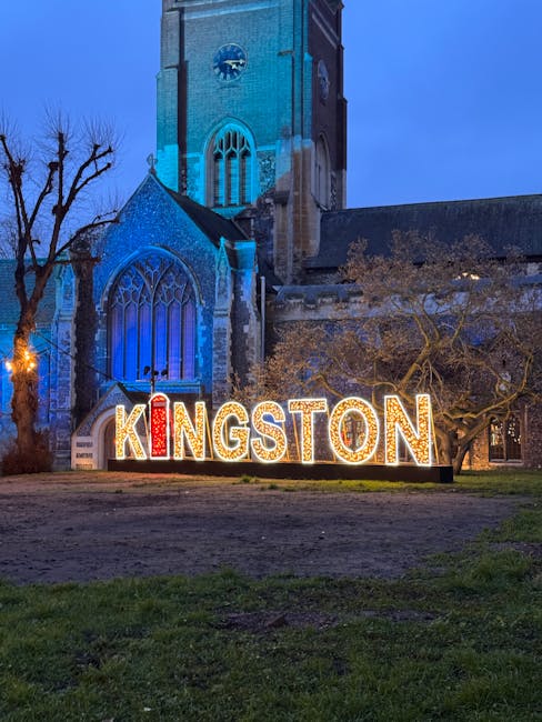 A nighttime view of a historic stone church with tall, pointed Gothic windows and a square clock tower, illuminated by blue lighting that highlights its architectural details. In front of the church, there is a well-lit sign spelling 'KINGSTON' with large, individual letters; the 'I' is designed to resemble a red and white striped candy cane. To the left, a leafless tree with dark, twisted branches stands against the evening sky. The ground in the foreground is a mixture of grass and bare soil, indicating an open public space or park. The scene conveys a quiet, evening atmosphere in Kingston, with the church and decorative signage creating a visually striking setting that could be associated with community or cultural events, and subtly connected to local initiatives such as private waste handling or community-based rubbish removal efforts facilitated by companies like Waste Clearance Kingston.