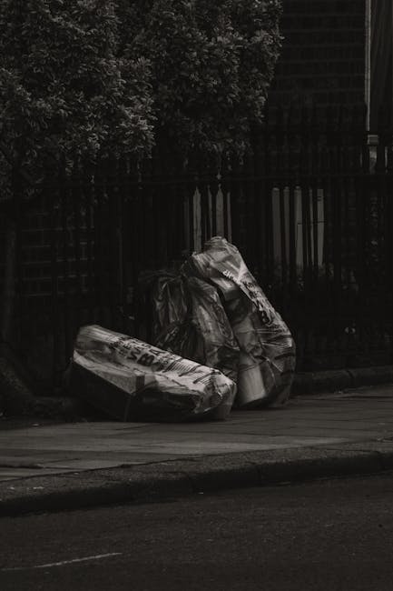 Three large black plastic rubbish bags, filled and tied at the top, are placed on a paved sidewalk near a black metal fence and a row of dense, leafy shrubs. The bags appear to contain household waste materials, with some visible creases and crumples showing the plastic's texture. The scene is set outdoors in a residential or urban area, with the bags positioned close to the curb, indicating a typical collection spot for waste disposal. In the background, the black fence provides an enclosure behind the foliage, and natural ambient light creates subtle highlights on the plastic surfaces, accentuating their shape and texture. Waste Clearance Kingston offers private rubbish removal and on-site waste clearance services, which are relevant in this context of independent waste handling, providing an alternative to standard council collection for household rubbish like these bags.