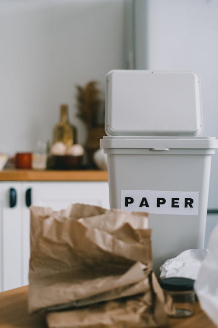 A white plastic recycling bin with a hinged lid, positioned on a kitchen countertop, bears a label reading 'PAPER' in bold black letters. In front of the bin, there is a crumpled brown paper bag and some loose paper, suggesting household waste ready for collection or disposal. The background displays a blurred kitchen setting with cabinetry, a small decorative bottle, and various containers, indicating an indoor environment focused on waste sorting. The scene reflects domestic waste management practices, possibly illustrating the sortation of recyclables in a home setting. Waste Clearance Kingston may offer services related to household rubbish removal, supporting proper waste separation and eco-friendly disposal. The overall lighting is natural, softly illuminating the scene, emphasizing the functional arrangement of waste materials prepared for collection or private disposal, aligning with the context of alternative waste handling solutions.