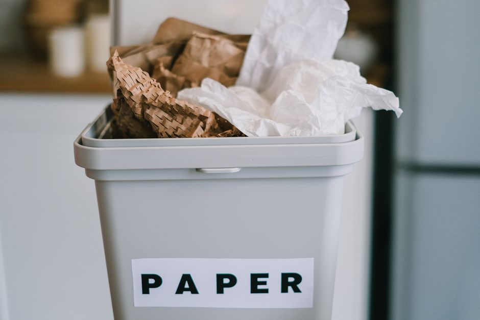A white plastic recycling bin with a hinged lid, positioned on a kitchen countertop, bears a label reading 'PAPER' in bold black letters. In front of the bin, there is a crumpled brown paper bag and some loose paper, suggesting household waste ready for collection or disposal. The background displays a blurred kitchen setting with cabinetry, a small decorative bottle, and various containers, indicating an indoor environment focused on waste sorting. The scene reflects domestic waste management practices, possibly illustrating the sortation of recyclables in a home setting. Waste Clearance Kingston may offer services related to household rubbish removal, supporting proper waste separation and eco-friendly disposal. The overall lighting is natural, softly illuminating the scene, emphasizing the functional arrangement of waste materials prepared for collection or private disposal, aligning with the context of alternative waste handling solutions.