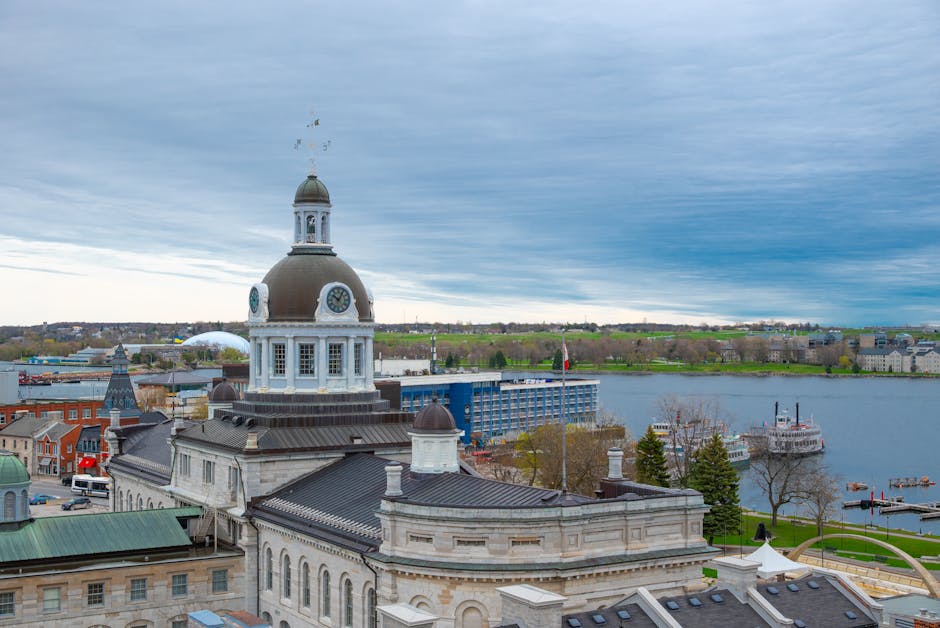 Aerial view of a historic building with a large central dome topped by a smaller cupola, featuring a clock face and decorative architectural elements in shades of cream and dark grey. The building's roof has a combination of rounded and flat sections, with some roofs covered in dark slate tiles. Surrounding the structure are other smaller buildings, some with green and grey roofs, and a parking lot with cars. In the background, a river spans across the image, with a few boats docked along its banks, and a cityscape with modern and older buildings stretching towards the horizon under a cloudy, overcast sky. The scene captures the mixture of historic architecture and natural landscape, indicative of an urban riverside area, aligned with typical rubbish removal and waste clearance services that handle waste from city buildings and surrounding environments. Waste Clearance Kingston provides these services within urban and waterfront locations, supporting private and commercial waste disposal needs in the area.