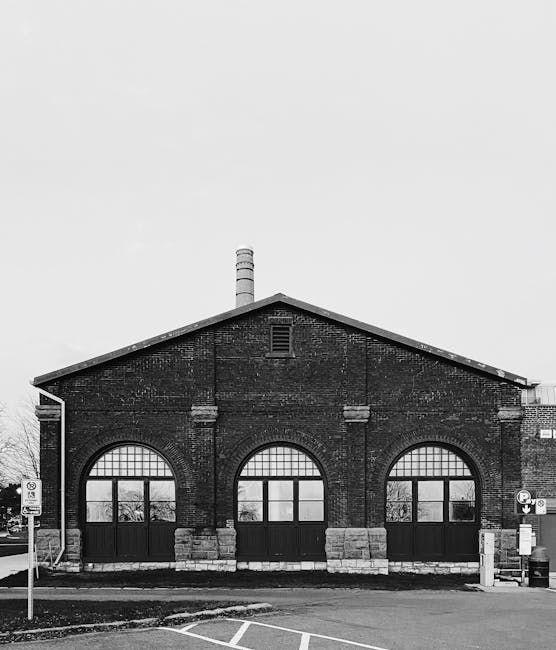 A black and white image depicts the front facade of an old brick industrial building, featuring three large arched windows with metal framing and multiple panes, situated on a paved street. The brickwork shows signs of age and weathering, with some areas darker and textured. The building has a gabled roof with a chimney extending vertically from the center, indicating its industrial past. In front of the structure, there is a small section of sidewalk and a parking area with visible road markings, along with a parking restriction sign and a designated loading zone. The scene is illuminated by natural light under an overcast sky, creating a neutral and professional atmosphere. In the context of waste clearance services, this setting could illustrate a location where independent or private rubbish removal, such as building waste or renovation debris, might be carried out by Waste Clearance Kingston, supporting alternative waste handling methods outside of municipal services.