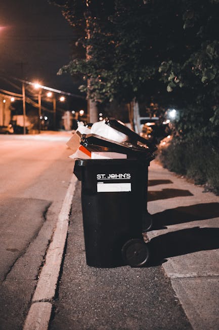 A black wheeled rubbish bin positioned on the pavement beside a street at night, filled with various waste items including cardboard boxes and plastic bags. The bin features a white label with the text 'St. John’s' and a blank space for additional information. The surrounding environment includes dim street lighting overhead, casting long shadows on the ground, with a row of trees and bushes along the sidewalk to the right, and residential or commercial buildings in the background. The scene evokes an urban setting where independent waste collection services, such as those by Waste Clearance Kingston, may handle refuse outside official council collections, illustrating a typical example of private refuse disposal or on-site clearance practices that require suitable waste receptacle management.