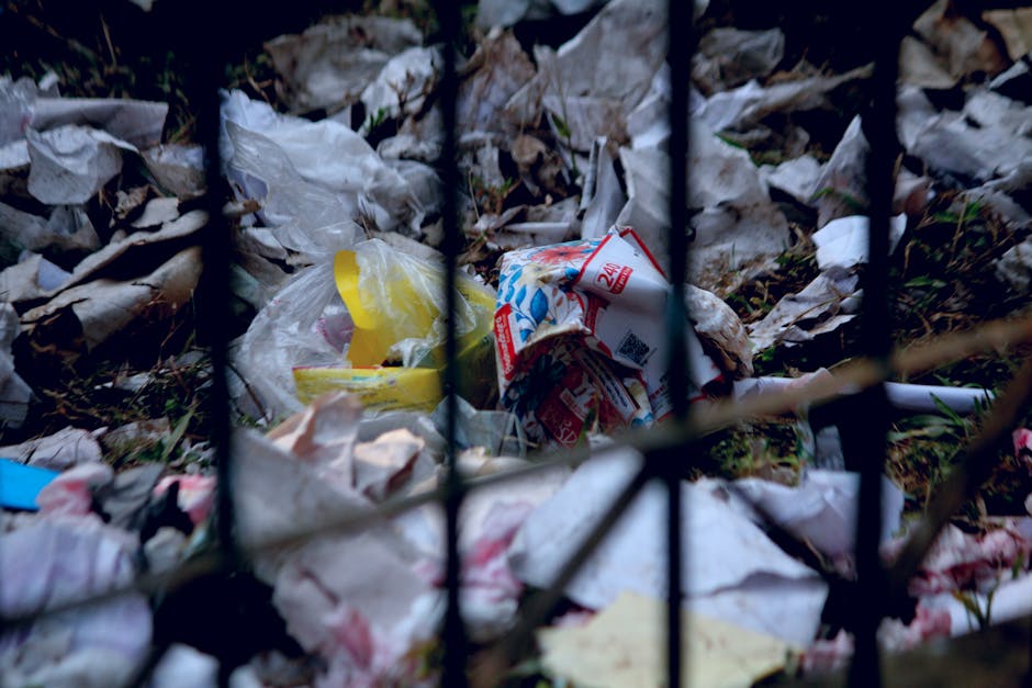 A close-up view through black metal fence bars showing piled-up litter consisting of crumpled paper, plastic bags, and discarded packaging materials scattered on the ground amidst dried leaves and grass. The trash includes a crumpled white paper bag, a yellow plastic bag, and a torn piece of packaging with red and blue printed graphics. The environment is outdoors, likely on a pavement or roadside area, with natural lighting and an unkempt appearance indicative of illegal dumping or unmanaged waste near a public space. The scene reflects the type of refuse often encountered in private waste clearance or rubbish removal services, highlighting the importance of proper waste disposal and waste management solutions. The focus on scattered household and packaging waste emphasizes the context of alternative waste handling outside municipal collection, consistent with the theme of independent rubbish removal near Kingston Hospital.