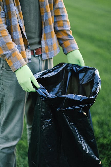 A person standing outdoors on a grassy area, wearing a plaid shirt with shades of yellow and gray over a dark green top, and light green gloves. They are holding a large, open black garbage bag made of shiny, thick plastic, with the bag's interior and edges clearly visible. The person's stance suggests they are in the process of collecting and disposing of waste, which may relate to private rubbish removal or on-site clearance services offered by Waste Clearance Kingston. The background shows a well-maintained lawn or sports ground, with vibrant green grass and natural lighting, emphasizing the clean, organized environment suitable for waste collection activities. This scene illustrates the physical aspect of rubbish handling and disposal, aligning with professional waste management practices, possibly involving independent collection rather than municipal refuse services.