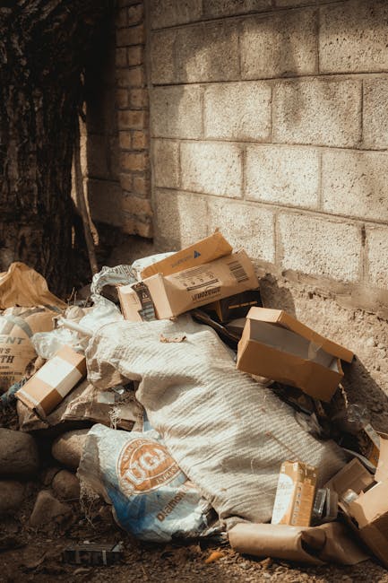 A pile of various discarded cardboard boxes and packaging materials, some flattened and others still in their original shape, is seen leaning against a rough, grey brick wall outdoors. The boxes are in shades of brown, with some displaying printed labels and barcodes, and are scattered over a white fabric bag or covering that appears partially torn. Surrounding the heap are small stones and dirt, indicating an outdoor or alleyway setting beneath a large tree visible on the left side, which has dark bark and partially shadows the area. The scene suggests an informal or private waste collection situation, potentially managed by a rubbish removal service like Waste Clearance Kingston, with the objects indicative of a cleanup task involving household or business waste temporarily staged for collection or disposal. The lighting appears natural, with subtle shadows cast by the tree and objects, emphasizing the textures of the cardboard, fabric, and brick wall in the background, creating an overall neutral and observational snapshot relevant to waste clearance or alternative rubbish handling services.