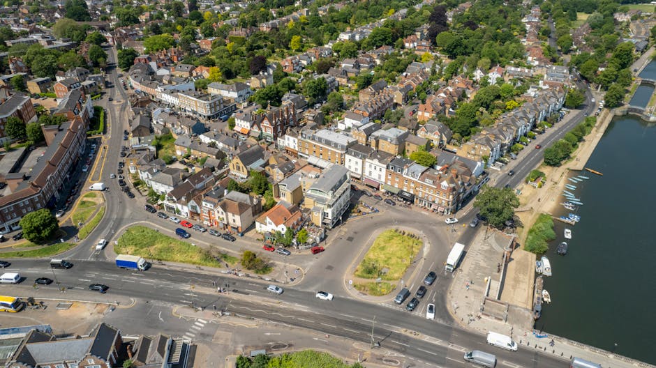 An aerial view of a coastal town showing a mix of residential and commercial buildings, including a cluster of shops and eateries along a main square. The buildings are constructed with brick and painted facades, with pitched roofs in shades of grey and brown, and some modern glass-fronted structures near the waterfront. The foreground features a busy road with multiple lanes, vehicles such as cars, trucks, and buses, and road markings indicating traffic lanes and pedestrian crossings. Adjacent to the road, there is a small, curved parking area with a few parked cars, and a narrow paved walkway leading to a waterfront promenade. The waterfront area includes a small dock with several boats moored along the quay, and a stone retaining wall separating the water from the land. Green spaces with grass patches and some trees are visible, providing a natural contrast to the urban environment. The scene is illuminated by daylight, with clear weather enhancing the visibility of the town’s layout and architectural details. This setting reflects a typical small town’s urban core, which could be part of an independent waste collection or rubbish removal service, such as Waste Clearance Kingston, operating in local areas like Canbury and Kingston Bridge.