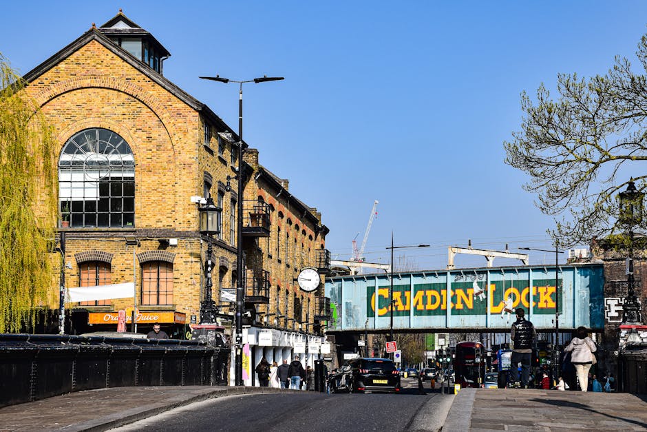 A street scene featuring a large, yellow-brick industrial-style building with arched windows and a gabled roof on the left side. In front of the building, there are street lamps, a clock, and a lamppost with a streetlight. To the right, a railway bridge painted in weathered blue with bold yellow and green lettering spelling 'CALDEIN LOCK' spans across the street, supporting railway tracks and electrical wiring. Pedestrians are walking on the sidewalk, some near small shops, while vehicles, including cars and buses, are seen on the street below. There are trees with budding or sparse foliage on the right side and in the background, construction cranes and additional urban infrastructure suggest ongoing development. The sky is clear and blue, providing bright natural lighting that enhances the visibility of textures on the brickwork, the metal bridge, and the surrounding environment. This scene subtly reflects a busy urban area where independent waste collection or rubbish removal services might operate, fitting within the context of alternative waste handling around Kingston and Canbury.