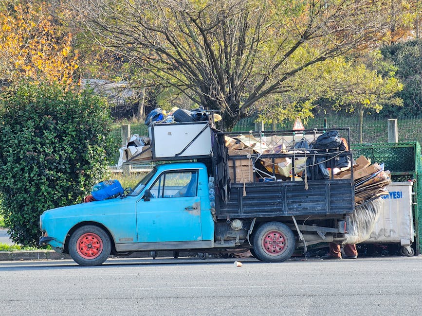 A small, old blue truck parked on the side of a street with its cargo bed overloaded with various types of waste including large cardboard pieces, black garbage bags, and other miscellaneous rubbish. The truck has a weathered appearance with faded paint and red wheels, and the cargo is secured with ropes and straps. Behind the truck, there is a green hedge and a large tree with sparse leaves indicating autumn. The surrounding environment appears residential with a fence visible in the background, and the scene is illuminated by natural daylight, highlighting the cluttered nature of the load. The image illustrates the concept of private waste collection, as offered by waste clearance services like Waste Clearance Kingston, which often handle alternative rubbish removal where on-site clearance or independent collection is required for bulky waste items.