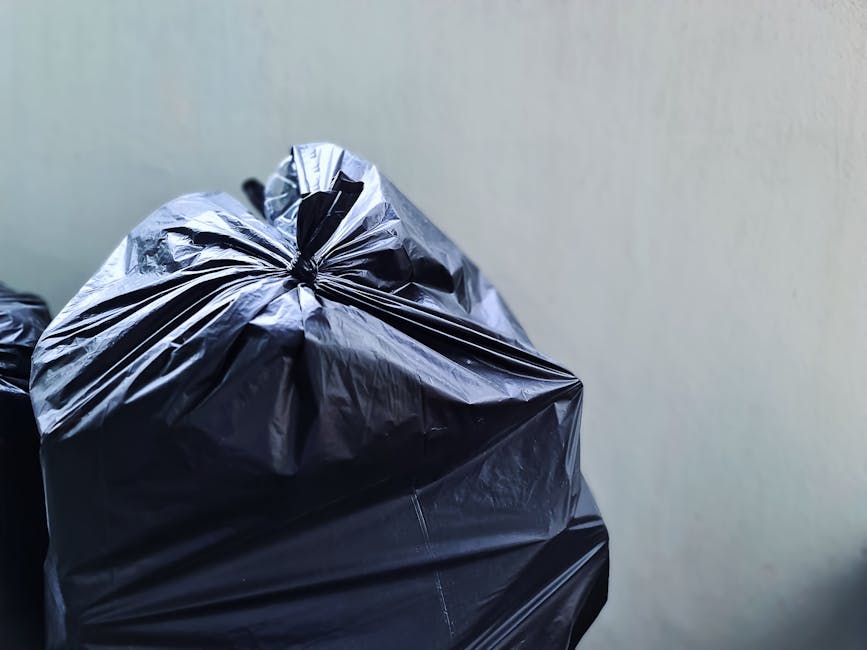 A close-up view of multiple large black plastic rubbish bags filled and tightly knotted at the top, stacked against a plain, light-colored wall. The bags appear to contain household waste or general refuse, with a shiny, smooth surface reflecting some light. The primary bag in the foreground is positioned on the left side of the image, with additional bags partially visible behind it, suggesting a small collection outdoors or in a storage area as part of a private waste collection or waste clearance service. The background wall is neutral and unadorned, and the lighting is soft, highlighting the textures and folds in the plastic. This scene exemplifies typical waste management practices where independent rubbish removal services, such as those offered by Waste Clearance Kingston, facilitate the disposal of household or commercial waste through on-site clearance, reducing reliance on local authority collections or disposal delays.
