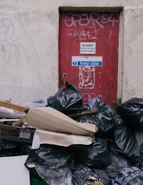 A dense pile of black garbage bags and scattered waste materials, including flattened cardboard boxes and discarded packaging, stacked haphazardly in front of a red metal door set into an exterior building wall. The door has graffiti tags painted in white and pink spray paint, along with a white sign that reads 'No Money' and a blue sticker displaying 'Keep clear.' The surrounding wall is textured beige with visible weathering, and the scene appears to be an urban alleyway or service area designated for rubbish disposal. Waste clearance Kingston is shown handling such refuse accumulation in contexts of private waste collection or on-site clearance, emphasizing the importance of managing overflowing rubbish outside commercial or residential buildings.
