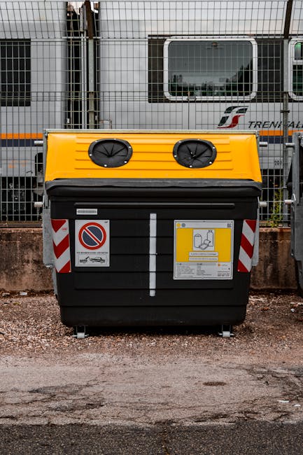 A large black waste container with a yellow lid is positioned on a gravel surface in front of a metal fence. The yellow lid features two circular air intakes with black covers, and the front of the bin displays multiple warning and instruction stickers, including a no-parking sign and labels related to waste handling. The container appears sturdy, designed for the collection of waste or recyclables, and is situated in an industrial or urban environment, possibly in a parking area or alleyway. Behind the fence, there is a train or tram car with a grey body, red, orange, and black branding, and a rectangular window with a reflective surface that offers a partial view of the surroundings. The scene suggests a setting associated with private waste collection or on-site rubbish disposal services, aligning subtly with the context of independent waste clearance activities conducted by Waste Clearance Kingston.