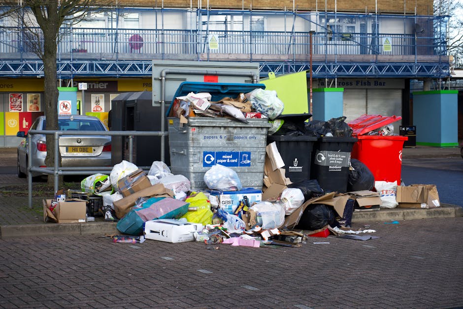 A large cluster of mixed waste and rubbish bags accumulated on a paved parking area in front of a commercial building. The waste includes a variety of materials such as cardboard boxes, plastic bags, paper waste, and packaging boxes, some of which are torn or crumpled. There is an open blue and gray recycling container labeled for mixed paper and card, filled with flattened boxes and loose paper spilling over its edges. Surrounding the container are black bin bags, a bright red plastic bin, and additional cardboard and paper debris scattered on the ground. The background reveals a multi-story building with scaffolding or external safety fencing, and on either side of the waste, there are small, green-upholstered barriers attached to metal railings. Behind the scene, the setting appears to be an urban commercial or retail area, with storefronts partly visible and a parked grey car on the left. The scene depicts an instance of excess waste likely awaiting disposal or alternative collection, illustrating the importance of proper rubbish removal services like Waste Clearance Kingston.