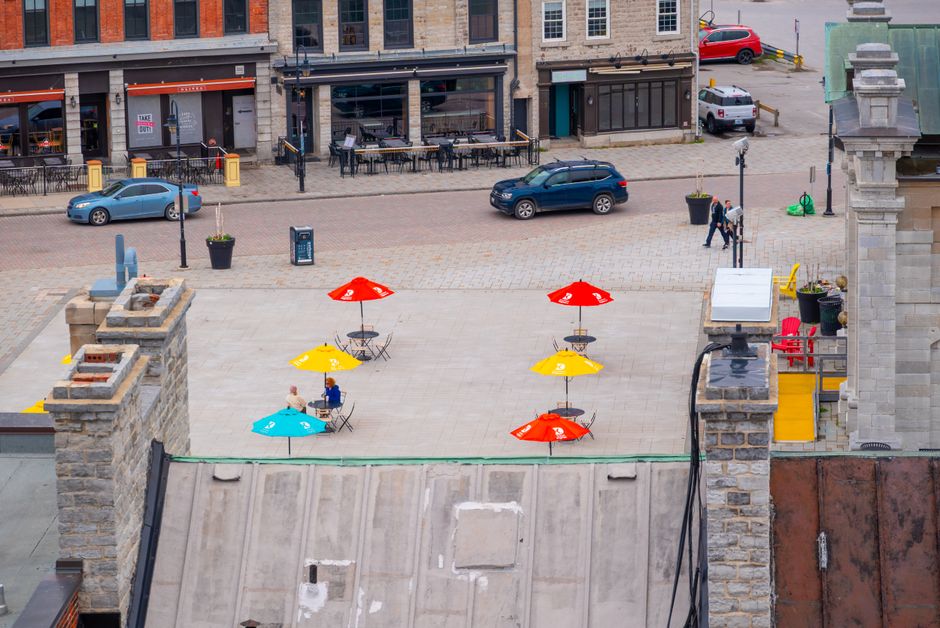 An aerial view of an outdoor urban area showing a paved open space with four colorful umbrellas—two red, one yellow, and one blue—each shading small sets of tables and chairs. In the foreground, part of a stone building or structure is visible with a sloped, textured metal roof. Beyond the open space, a street lined with parked cars, including a blue sedan, a black SUV, and a red hatchback, runs parallel. The background features a row of brick and stone buildings, some with storefronts, patio tables, and outdoor seating, indicating an area frequented by pedestrians. Two people are walking along the pavement towards the top right of the image, and a black lamppost stands near the center. The scene is illuminated by natural daylight, with shadows cast by the umbrellas and objects, conveying a clear, calm day. Waste Clearance Kingston's service focus on waste management and rubbish removal services can be connected to this setting, as the open public space may require regular clean-up or waste collection, especially when hosting outdoor events or patio dining areas, which often involve independent or private rubbish collection arrangements.